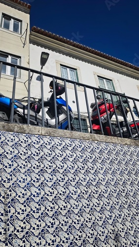 Motorcycles parked behind a decorative blue and white tiled wall with a railing, featuring a building facade and clear blue sky above