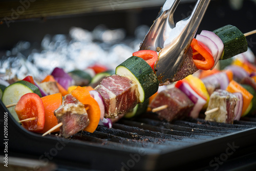 Colorful beef and vegetable kebabs sizzle over an outdoor barbecue grill as metal tongs turn the skewers for even cooking.