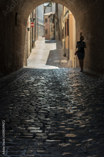 Sunlit cobblestone street framed by a shaded archway in an Italian town, with a lone pedestrian seeking relief from the summer heat. Selective focus.