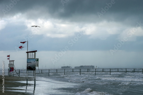 Storm clouds gather over an empty beach as red warning flags ban swimming, with rough waves and strong wind creating a dramatic coastal scene.