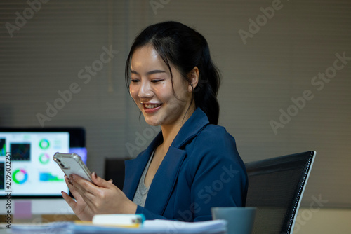 A happy Asian businesswoman smiles while scrolling through her smartphone during a late-night work break at the office.