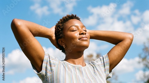 Serene young Black woman relaxing outdoors with eyes closed, enjoying sunshine and fresh air against a blue sky.