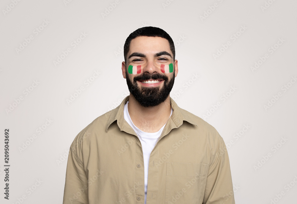 Fototapeta premium Happy Young Male, Middle Eastern, Celebrating National Pride With Italian Flag Face Paint. Posing Cheerfully In Studio Against Plain Background, Expressing Joy And Enthusiasm For Culture.