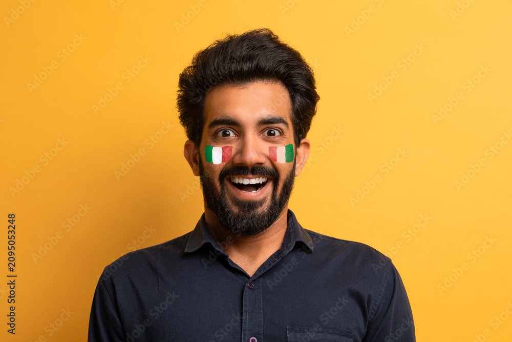 Fototapeta premium Smiling Indian Male With Face Paint In National Colors, Expressing Joy And Excitement In Studio Setting, Celebrating A Sports Event. Energetic Young Adult Man Showing Passion And Enthusiasm.
