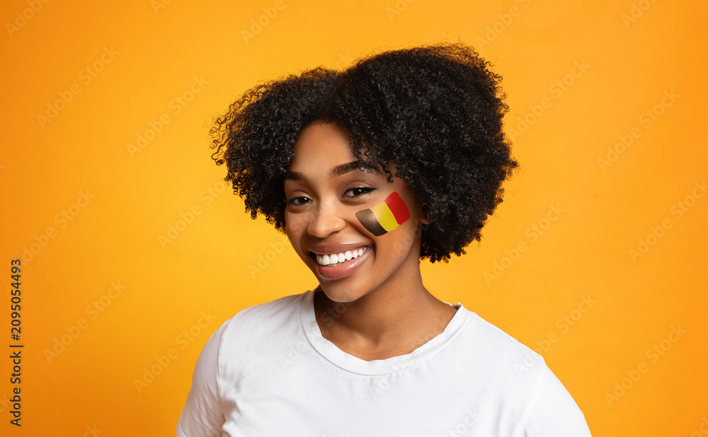 Fototapeta premium Happy Young Black Woman Smiling With Flag Painted On Face, Standing Against Yellow Background. Excited About Cultural Celebration In Studio. Represents Joy And Pride In Identity, Lifestyle Concept.