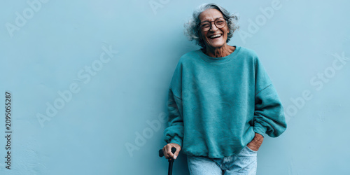 Smiling Senior Woman in Teal Sweatshirt, Holding Cane, Against Light Blue Wall - Portrait of Confidence and Joy