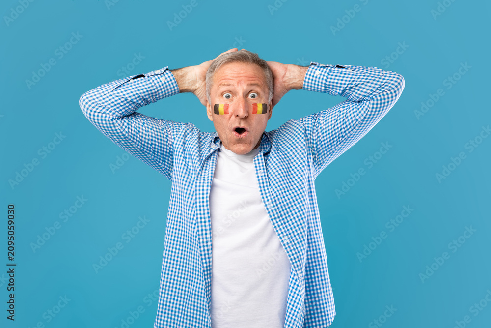 Fototapeta premium Surprised Elderly Caucasian Male With Face Paint Showing Excitement With Hands On His Head, Standing In Front Of Blue Studio Background, Celebrating Sports Event Or National Holiday.