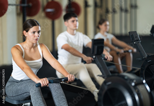 Workout girl cross training exercising cardio using rowing machine with group of people in crossfit club