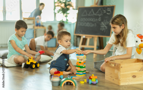 Boy and a girl of different ages sit together near a construction set and assemble it against the background of other children in kindergarten