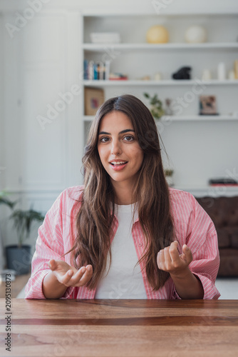 Fotografie Young woman speaks to camera while seated at a wooden desk in a bright living room, gesturing naturally during a casual video call or remote meeting, conveying confidence, warmth and approachability