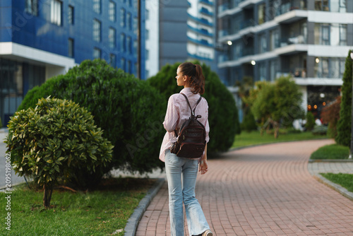 Konstfotografi Woman with backpack walking along a brick path in a modern residential district, showcasing casual lifestyle, urban exploration, and outdoor leisure
