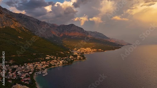 Aerial view of Brela Croatia shows the town by the sea. Mountains are visible in the background. Sunlight reflects on the water. Clouds fill the sky.
