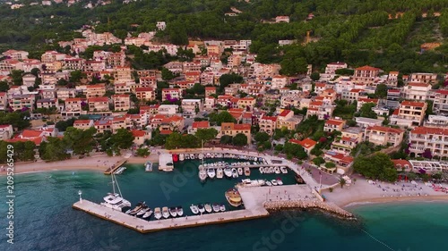 Brela features a coastal area with various residential buildings. Boats are docked in the marina. The beach area is visible. Green hills are in the background.