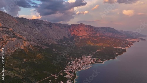 Brela shows an aerial view of the coastline and mountains. The village is near the water. Mountains are visible in the background. The sun is setting.