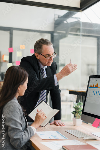 Business mentor explaining data charts on computer screen to female colleague in modern office