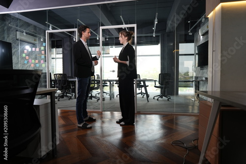 Photos A bearded professional and a bun-haired woman face each other for a standing talk beside a sticky-note glass wall in a warmly lit coworking office, an easygoing end-of-day conversation
