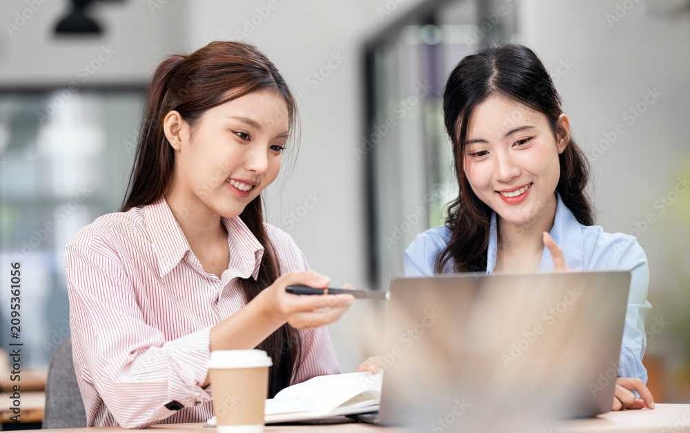 Obraz premium Two Smiling Young Asian Businesswomen Collaborating and Working on Laptop