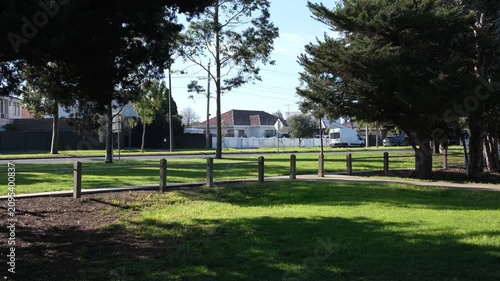 A quiet, shaded roadside green space, grassy reserve, or nature strip with a low fence separating road traffic and the footpath, residential houses, and running traffic in back. Melbourne, Australia