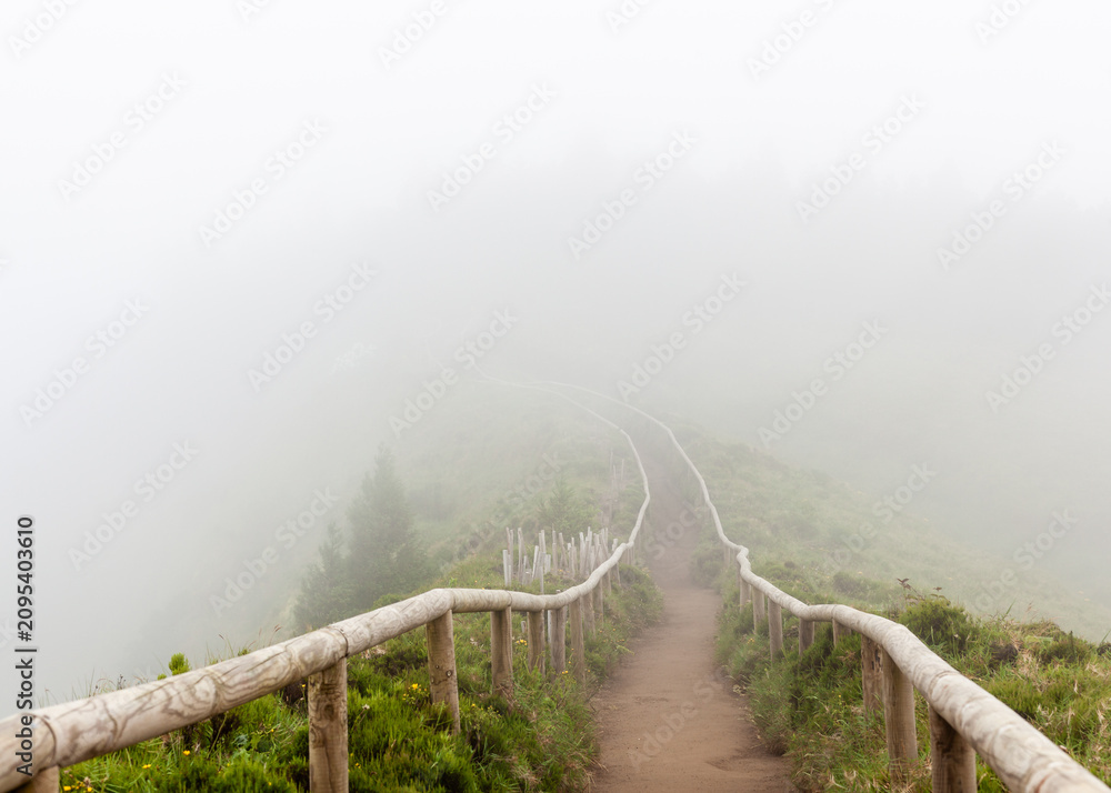 Fototapeta premium Misty hiking trail in the Azores. A wooden fence lines the path as fog obscures the landscape. Lush green vegetation covers the mountainside.