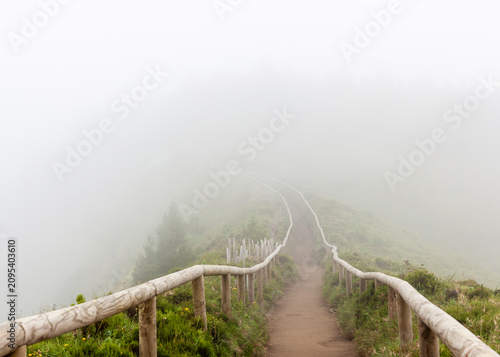 Misty hiking trail in the Azores. A wooden fence lines the path as fog obscures the landscape. Lush green vegetation covers the mountainside.
