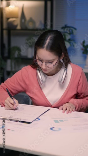 Vertical: Asian businesswoman wearing eyeglasses and wireless headphones around neck, holding pen to write on document at desk.