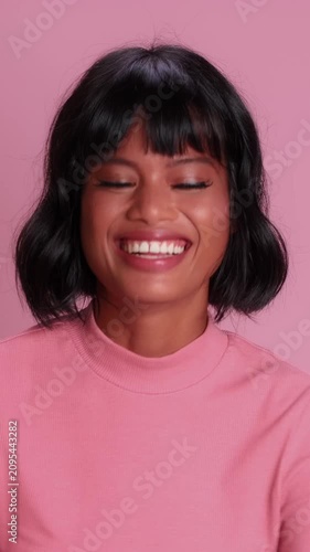 Hopeful Asian woman with short black hair crosses fingers on both hands, closes eyes tightly, and smiles warmly while wishing for good luck on solid bright pink studio background.