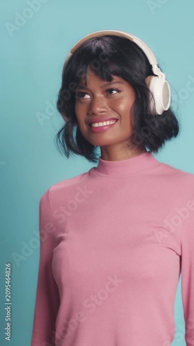 Smiling Asian woman with short black hair wears wireless over-ear headphones, nodding and enjoying favorite acoustic songs with beautiful warm smile on solid light blue studio background.