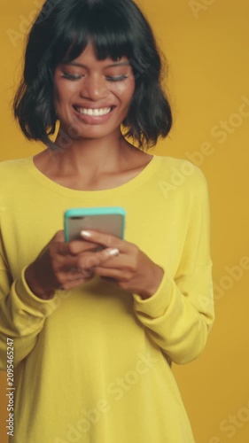 Smiling Asian woman with short black bob haircut types on touchscreen phone, looking down cheerfully, chatting online, and looking up to smile warmly on solid yellow studio background.