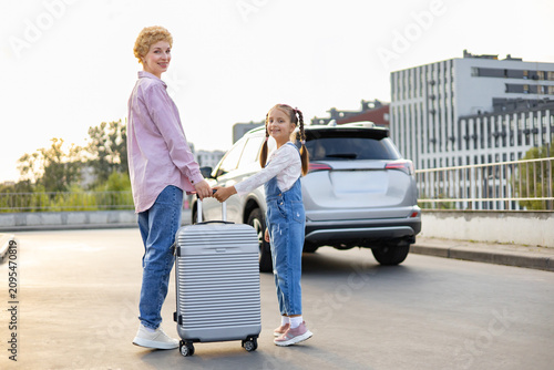 A Caucasian mother and her daughter stand with a suitcase next to a car, ready for a trip