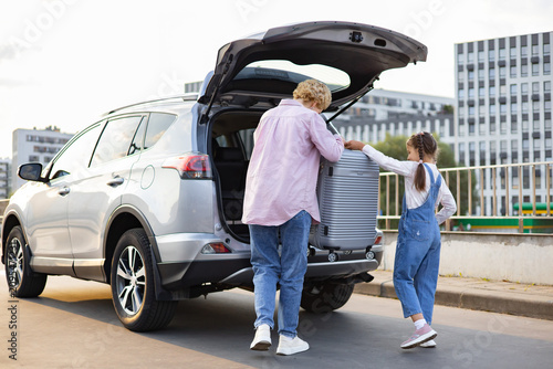 A Caucasian mother and her daughter are loading a suitcase into the trunk of a car, preparing for a journey