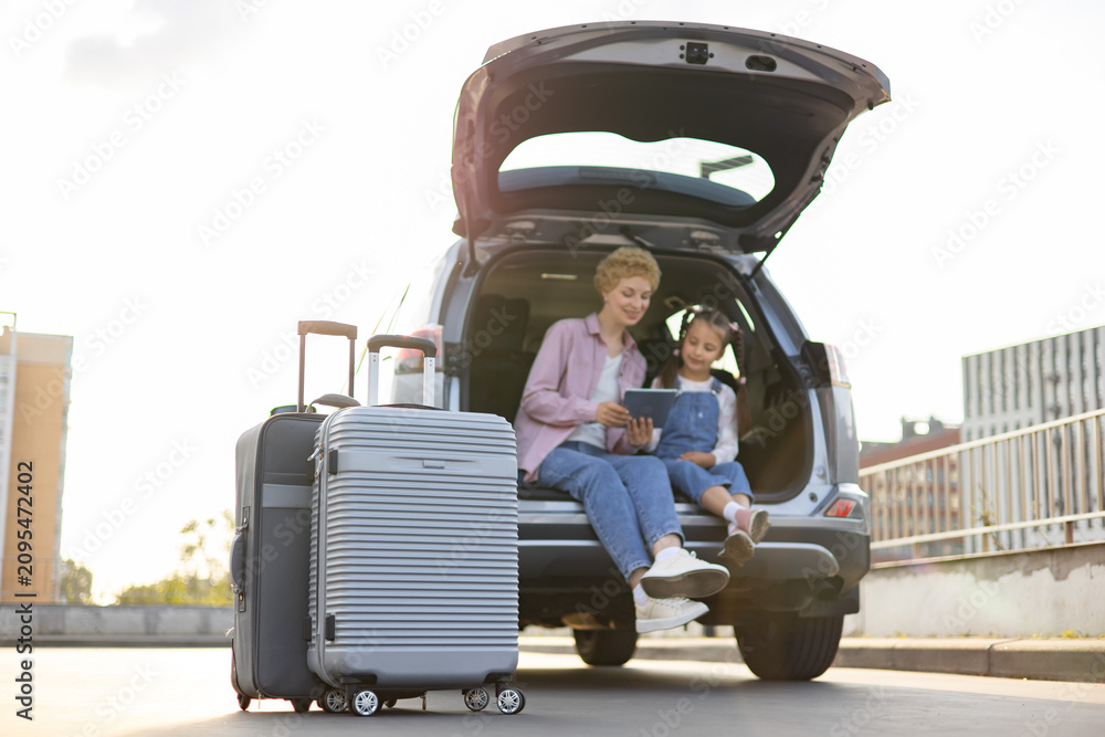 Obraz premium A Caucasian mother and her daughter are sitting in the open trunk of a car with luggage, looking at a tablet before their journey