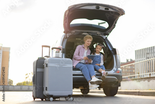 A Caucasian mother and her daughter are sitting in the open trunk of a car with luggage, looking at a tablet before their journey