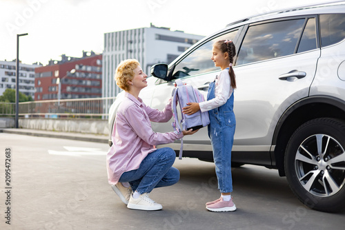 A mother hands a backpack to her daughter next to a car, preparing for school or an outing