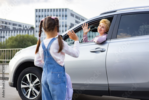 A smiling mother in a car waves goodbye to her daughter who is standing outside with a backpack