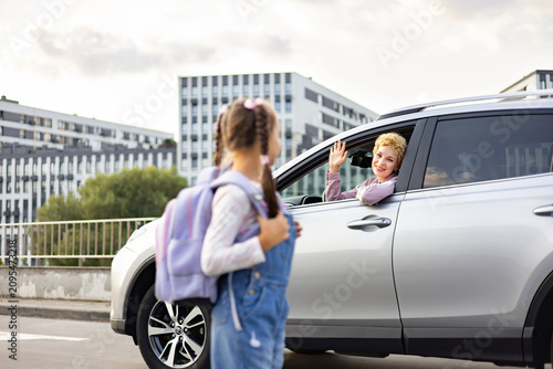A mother in a car waves goodbye to her daughter who is wearing a backpack and ready for school