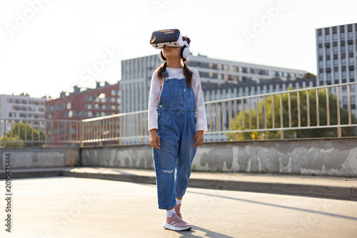 A young Caucasian girl wearing denim overalls and a VR headset explores a virtual world while standing on a rooftop with city buildings in the background