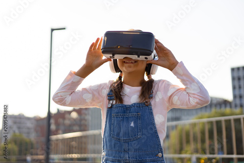 A young Caucasian girl wearing a VR headset and headphones is immersed in a virtual world outdoors