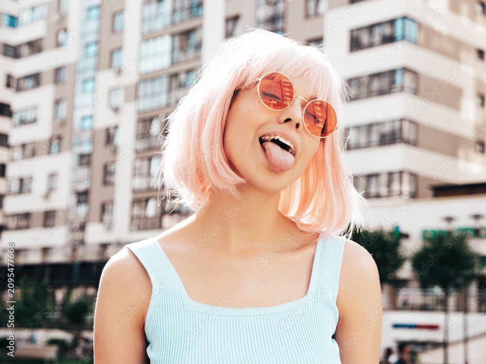 Fototapeta premium Young beautiful confident hipster female in trendy summer clothes. Sexy carefree woman posing in the street in wig. Positive funny model having at sunset. In sunglasses. At sunny day. Shows tongue