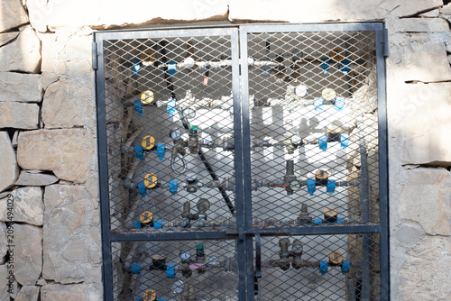 Metal mesh gate enclosing industrial water control valves mounted on a stone wall, showcasing plumbing infrastructure and maintenance access