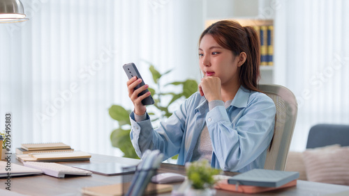 Young Asian student using smartphone for online education while studying at desk in bright home workspace