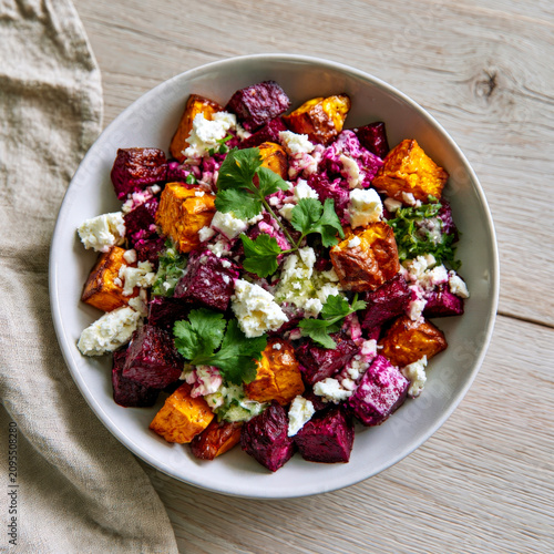 Roasted beetroot and sweet potato salad with crumbled feta cheese and fresh cilantro in white bowl on wooden table