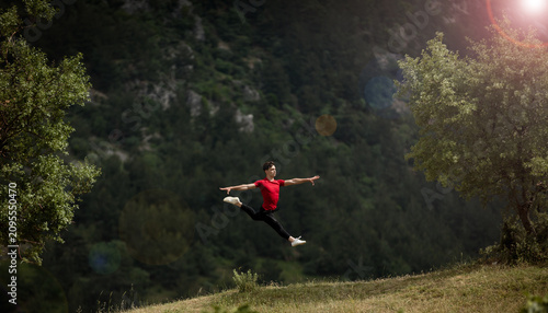 Quadro su tela Spectacular mid-air split jump of a young male dancer or athlete in a red t-shirt against a lush green forest and mountain background