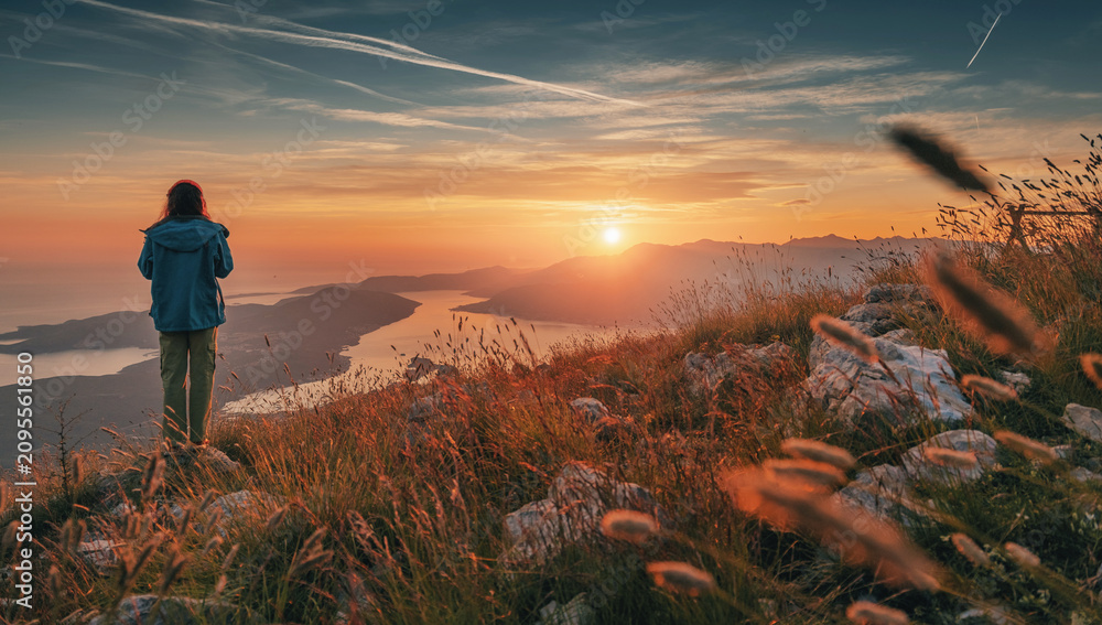Obraz premium Woman standing on mountain top at sunset, looking across scenic bay and mountains, finding freedom and peace in nature