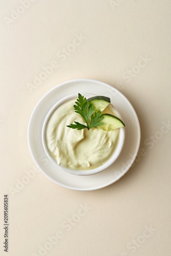 Creamy hummus dip bowl with cucumber slices and parsley garnish on a beige background. Top view, negative space