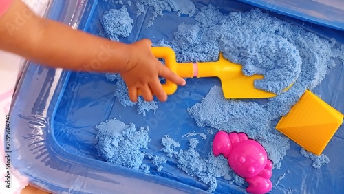 Photos Child's hand playing with blue kinetic sand in a plastic tray using yellow shovel and pink crab mold