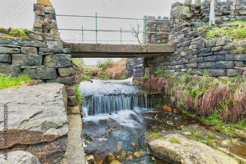 A stone bridge spans a small river with a tiered waterfall, bordered by historic stone walls and lush greenery, suggesting a scenic countryside location - Cheesden Lumb Mill, Lancashire, UK