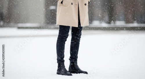 Person wearing a beige trench coat, dark jeans, and black chelsea boots in snow