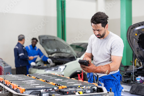 Auto technician testing electric vehicle battery modules in a service garage, with car hood open and team working in the background, focusing on diagnostics and maintenance.