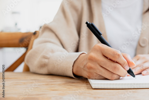 Close up of a person writing in a notebook with a pen on a wooden desk. Ideas for note taking, studying, planning and productivity concepts.