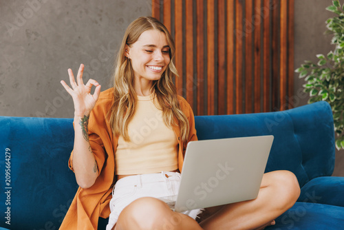 Young IT woman wears brown shirt hold use work on laptop pc computer sit on blue sofa couch stay at home hotel flat relax spend free spare time in living room indoors grey wall. People lounge concept.